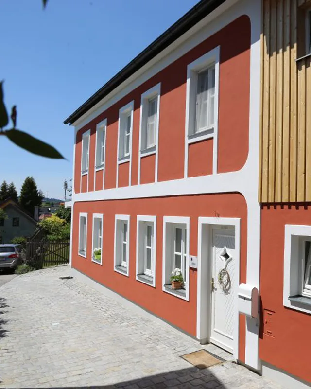 Zweistöckiges Haus mit roter Fassade und weißen Fensterrahmen in sonniger Wohngegend, mit gepflastertem Vorplatz und dekorierter Haustür.