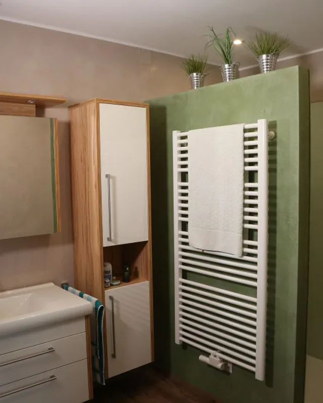 Modernes Badezimmer mit grüner Akzentwand, weißem Handtuchheizkörper und minimalistischer Holzmöblierung. Drei Zierpflanzen in Metalltöpfen setzen einen frischen Farbakzent.